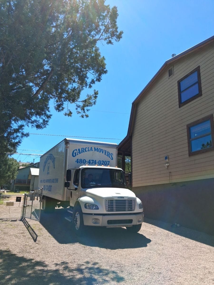 A Garcia Movers truck backed onto a gravel drive beside a two-story home under tall pines
