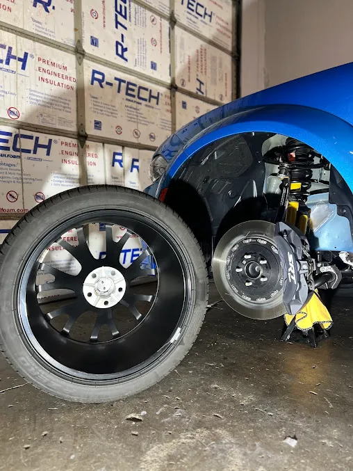 Black alloy wheel beside a blue sports car with exposed brake disc and yellow caliper